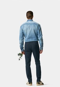 Veste en denim bleu clair au design ajusté, pantalon marine foncé, tenant une branche de verdure, et portant des baskets à motifs.
