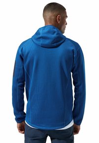 Blaues Kapuzensweatshirt aus strukturiertem Material, mit einem kontrastierenden schwarzen Patch am linken Arm und sichtbaren Nahtdetails.