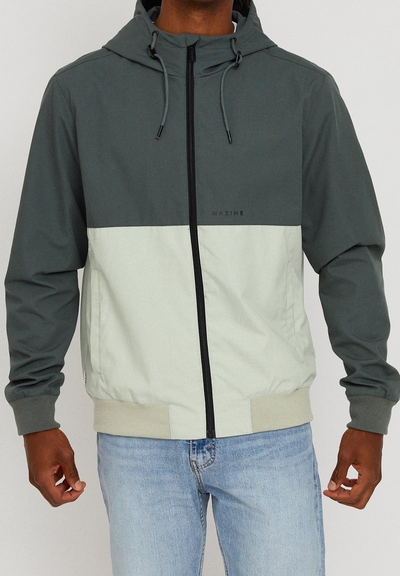 Homme portant une veste zippée à capuche bicolore gris et blanc cassé avec des poignets côtelés et un jean bleu clair.