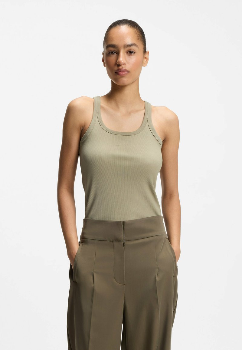 Jeune femme aux cheveux attachés portant un débardeur beige sans manches et un pantalon taille haute vert olive avec les mains dans les poches.
