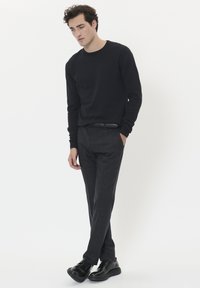 Svart långärmad skjorta i kombination med grå rutiga byxor; modell har svarta skor på sig, med händerna i fickorna, står mot en vit bakgrund.