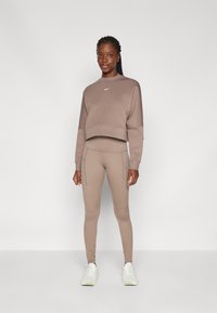 Felpa corta marrone con un piccolo logo, abbinata a leggings coordinati con tasche laterali e una vestibilità slim. Footwear sportivi bianchi.