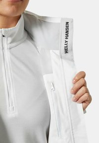 Chaqueta blanca de Helly Hansen con cremallera frontal, textura suave, cuello alto y logo en el cuello. Cuenta con un pequeño bolsillo con cremallera en el lado.