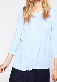 Blusa azzurra chiara con maniche a tre quarti, caratterizzata da pieghe verticali, un collo arrotondato e una vestibilità fluida. Tessuto morbido.