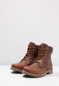 Botas de ante marrón hasta el tobillo con ojales de metal, cuello forrado de piel sintética y suela de goma marrón con un patrón de huella texturizado.