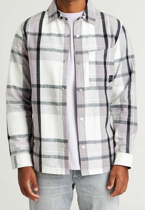 Uomo che indossa una camicia a quadri bianca e grigia sopra una maglietta bianca semplice e jeans grigio chiaro, in piedi davanti a uno sfondo neutro.