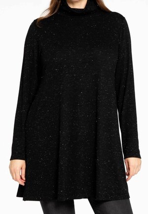 Femme portant un haut tunique noir tacheté à manches longues avec un col montant et une coupe ample sur un pantalon sombre.