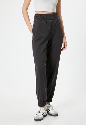 Black joggers with a smocked waistband, cuffed ankles, and side pockets, made of smooth fabric, paired with white and grey sneakers.
