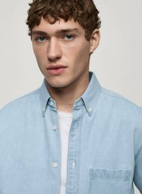 Camicetta di denim azzurro chiaro con colletto a bottoni, taschino frontale e dettagli di cucitura discreti, indossata sopra una t-shirt bianca.