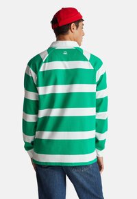 Długorękawowa koszula w zielono-białe paski z kołnierzykiem, z raglanowymi rękawami i małym logo na plecach. Połączona z niebieskimi dżinsami.
