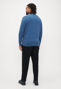 Niebieski sweter z dekoltem w serek i prążkowanymi mankietami, w zestawie z czarnymi spodniami i czarnymi butami, widziany od tyłu na jednolitym tle.