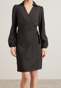 Robe noire cintrée avec un col, de longues manches bouffantes et un détail de taille nouée. Fabriquée dans un tissu lisse avec un design drapé à l'avant et une silhouette flatteuse.