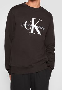 Černá mikina s výrazným bílým logem Calvin Klein Jeans. Elastické manžety a lem, klasický kulatý výstřih. Měkká textura látky.