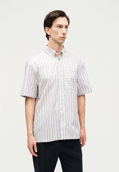 Άνδρας που φοράει κοντομάνικο πουκάμισο με κάθετες μπλε και πορτοκαλί ρίγες, στέκεται μπροστά σε απλό φωτεινό φόντο.