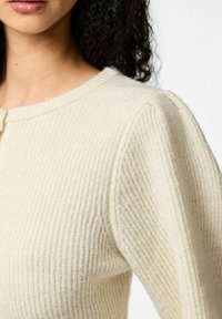 Flødefarvet ribstrikket sweater med rund hals, tekstureret strikket stof, diskrete pufærmer og en central knappedetalje.