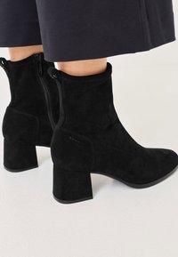 Crne nubuk čizme do gležnja s blok potpeticom, sa bočnim zatvaračem i čistim linijama. Tekstura je meka i glatka bez vidljivih uzoraka.