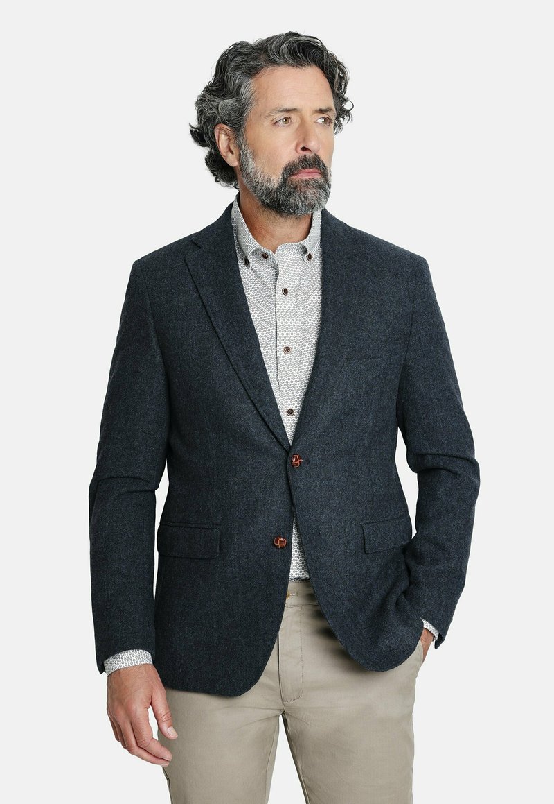 Blazer de lana navy con solapas notch, dos bolsillos frontales y botones marrones, usado sobre una camisa blanca con estampado, combinado con pantalones beiges.