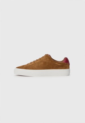 Brązowy niski zamszowy sneaker z białą podeszwą i czerwonym paskiem na pięcie, widziany z boku na jednolitym jasnym tle.