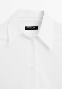 Gros plan sur le col d'une chemise blanche et les boutons supérieurs avec une étiquette noire indiquant "Massimo Dutti, Made in Portugal."