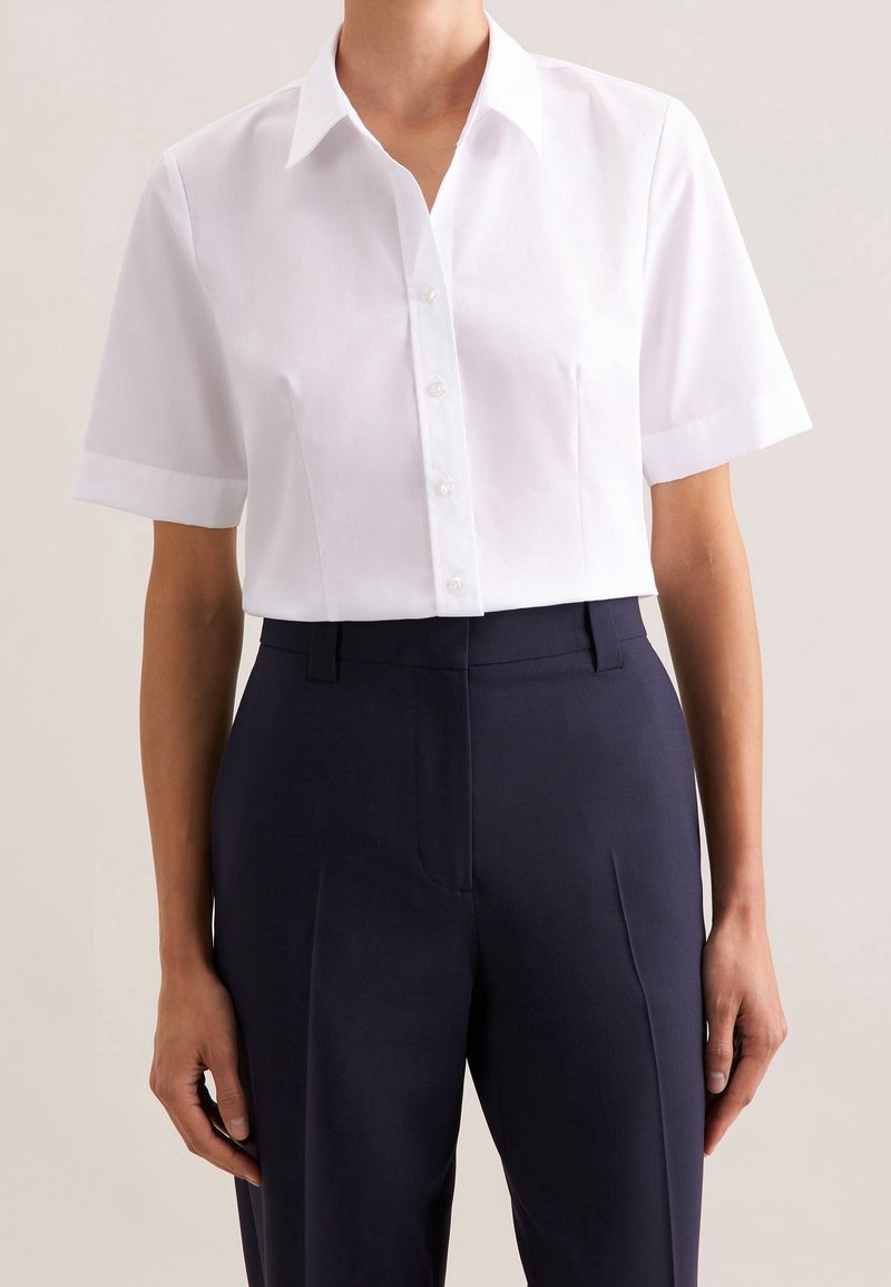 Personne portant une chemise blanche cintrée à manches courtes boutonnée, rentrée dans un pantalon taille haute bleu marine foncé, debout devant un fond uni.