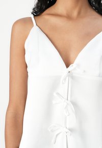 Top bianco senza maniche con profondo scollo a V, dettagli legati lungo il centro e una texture liscia e opaca.