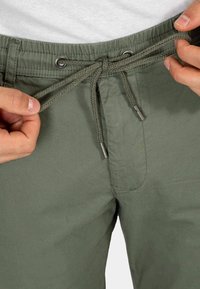 Pantalones verde oliva con cintura ajustable mediante cordón, que incluyen ojetes metálicos y cordones trenzados. Tela suave con diseño frontal plano.