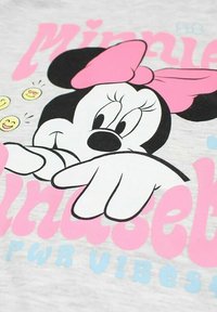 T-shirt gris avec un personnage de dessin animé, une souris noir et blanc avec un nœud rose, du texte en rose et bleu, et des icônes de visages souriants autour.