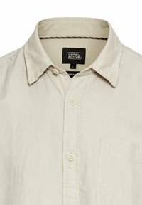 Beige linnen shirt met een kraag, twee knopen en een klein zakje; heeft een subtiele textuur en stikseldetails. Merktag zichtbaar aan de binnenkant.