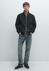 Black zip-up jacket with a high collar, worn over a gray shirt, paired with light blue straight-leg jeans and dark brown suede shoes.