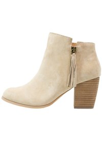 Beige suède enkelboot met een ronde neus, een zijrits en een kwastje. Heeft een ribbelige, gestapelde houten hak. Glad materiaal en minimalistisch design.