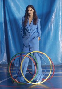 Completo su misura blu con giacca doppiopetto e pantaloni a gamba larga, abbinato a scarpe bianche, che si trova dietro a cerchi colorati su un campo.