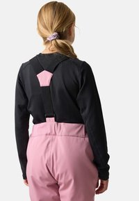 Lyserøde vandtætte overalls med justerbare sorte seler og en blød tekstur, kombineret med en sort bluse med lange ærmer, set fra bagsiden.