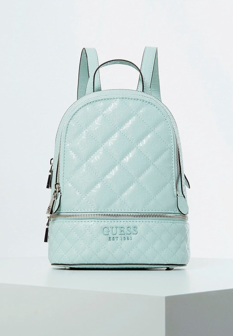 Sac à dos en simili cuir matelassé bleu clair avec une poche zippée à l'avant, une poignée supérieure et des bretelles réglables. Présente le logo "GUESS EST 1981".