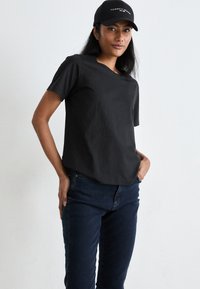 Black cotton t-shirt with a relaxed fit, crew neckline, and short sleeves paired with dark denim jeans and a black cap featuring white logo text.