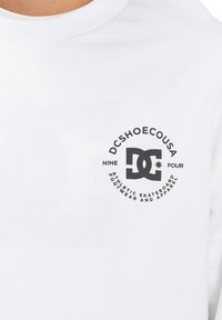 White long-sleeve shirt featuring a circular black logo with text "DCSHOECOUSA," "NINE FOUR," and "ATHLETIC SKATEBOARD FOOTWEAR AND APPAREL."
