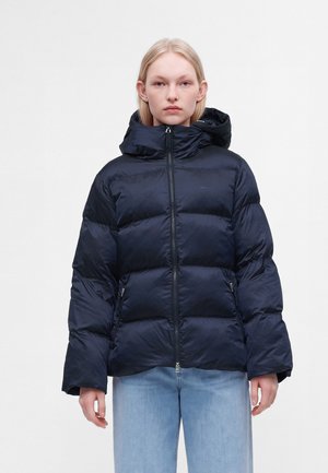 Jeune femme portant une doudoune bleu foncé avec capuche et un jean bleu clair, debout devant un fond uni clair.