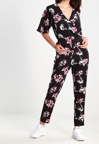 Jumpsuit noir à motifs floraux avec un design enroulé, présentant des fleurs roses et blanches, des manches larges et des jambes cintrées. Associé à des baskets blanches.