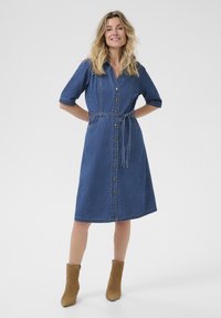 Denim obleka s kratkimi rokavi, gumbom ob sprednji strani, pasom na vezanje v pasu in dolžino do kolen. Izdelana iz modrega denima z gladko površino.