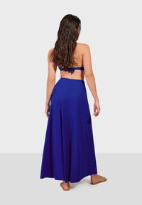 Modrá sukňa po zem s voľným strihom, vyrobená z ľahkej tkaniny. Má priliehavý pás s otvoreným chrbtom a nastaviteľnými ramienkami.