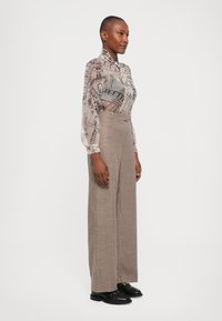 Transparante blouse met knopen en een architectonische print in grijs en beige, gecombineerd met hoog-waardige, geruite, wijdvallende broek in bruin en zwart.