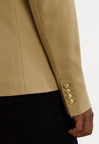 Manica di blazer beige texturizzato con dettagli di bottoni dorati, presenta una vestibilità tailored e cuciture visibili, abbinato a un capo nero.