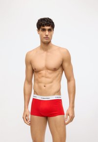 Czerwone bawełniane bokserki z białym elastycznym pasem, na którym widnieje nazwa marki „Calvin Klein”. Model stoi w neutralnej pozzie.
