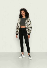 Kamuflāžas bomber jaka ar melniem akcentiem, pelēks īsais tops, melnas šaurās bikses un baltas kedas. Modelis stāv pret gaišu fonu.