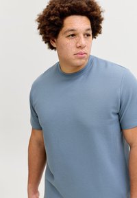 Νεαρός με σγουρά μαλλιά και φακίδ ες που φοράει ανοιχτό μπλε πουκάμισο με υφή και κοντά μανίκια, κοιτάζει προς τα δεξιά του μπροστά σε απλό φόντο.