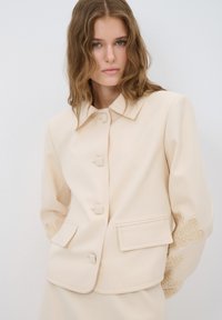 Beige gestructureerde jas met een puntige kraag, zeven met stof bedekte knopen, zijzakken en geborduurde accenten op de mouwen.