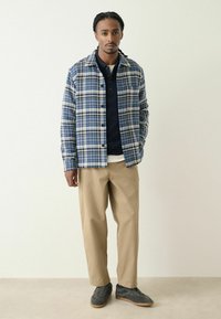Checkered blue and white shirt jacket over a navy polo, paired with beige trousers and grey sneakers. Smooth, lightweight fabric.