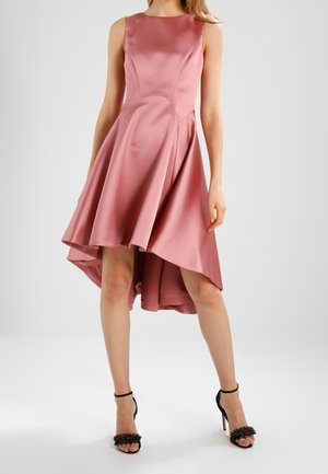 Femme portant une robe sans manches en satin rose à ourlet haut-bas et des sandales noires à talons hauts avec brides à la cheville et détails décoratifs.