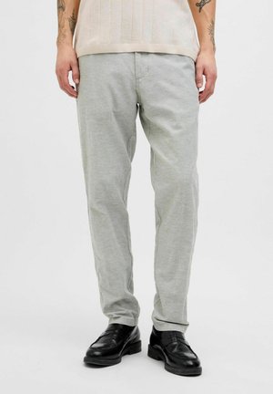 Homme portant un pantalon décontracté gris clair et des chaussures en cuir noires sans lacets, debout devant un fond blanc uni.