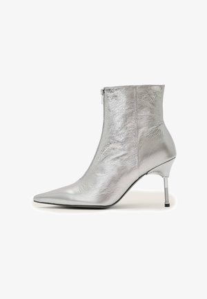 Zilverkleurige metallic enkellaars met een spitse neus, ritssluiting aan de zijkant en een slanke naaldhak met zwarte punt op een witte achtergrond.