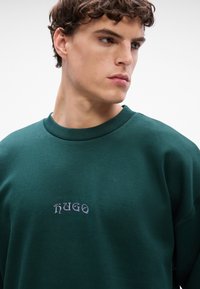 Tamnozeleni dukserica s okruglim izrezom i rebrastim detaljima. Na prsima se nalazi izvezena oznaka "HUGO" u svijetlo sivoj boji. Tkanina je u kombinaciji pamuka.